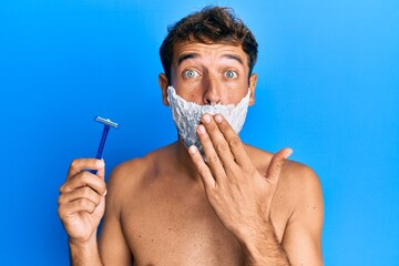 Handsome man saving beard with shave foam over face holding razor covering mouth with hand, shocked...