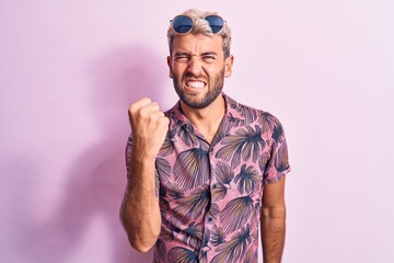 Naklejka premium Handsome blond man on vacation wearing casual shirt and sunglasses over pink background angry and mad raising fist frustrated and furious while shouting with anger. Rage and aggressive concept.