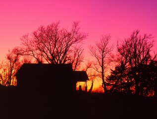 Silhouette of farmhouse and bare trees at sunset