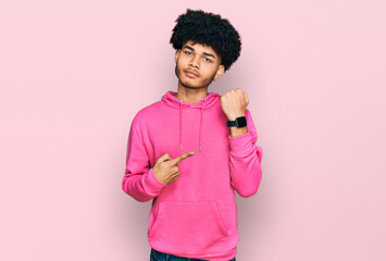 Young african american man with afro hair wearing casual pink sweatshirt in hurry pointing to watch...