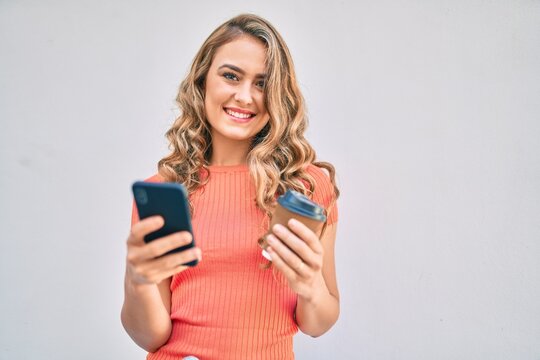 Young blonde girl smiling happy using smartphone and drinking take away coffee at the city.