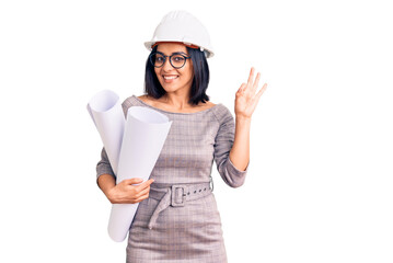 Young beautiful latin woman wearing architect hardhat and glasses holding blueprints doing ok sign with fingers, smiling friendly gesturing excellent symbol