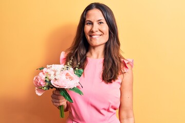 Young beautiful brunette woman holding bouquet of pink flowers over yellow background looking positive and happy standing and smiling with a confident smile showing teeth