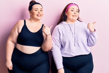 Young plus size twins wearing sportswear smiling with happy face looking and pointing to the side with thumb up.