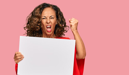 Middle age hispanic woman holding blank empty banner annoyed and frustrated shouting with anger, yelling crazy with anger and hand raised