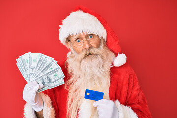 Old senior man wearing santa claus costume holding dollars and credit card in shock face, looking skeptical and sarcastic, surprised with open mouth