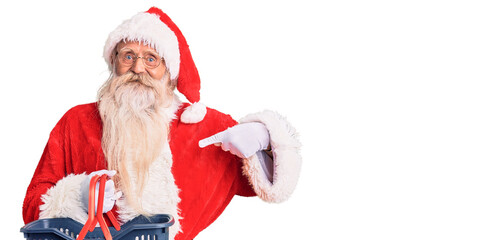Old senior man with grey hair and long beard wearing santa claus costume holding shopping basket pointing finger to one self smiling happy and proud