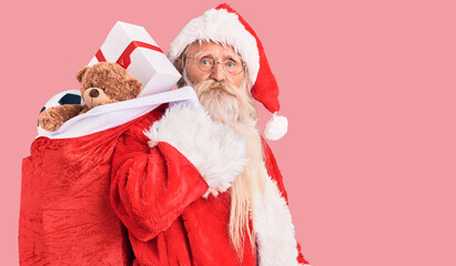 Old senior man with grey hair and long beard wearing santa claus costume holding bag with presents thinking attitude and sober expression looking self confident