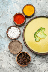 Vertical view of creamy broccoli soup in a brown bowl and different spices on gray background
