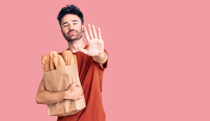 Young hispanic man holding paper bag with bread with open hand doing stop sign with serious and...