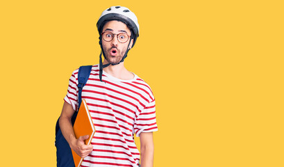 Young hispanic man wearing student backpack and bike helmet holding binder scared and amazed with open mouth for surprise, disbelief face