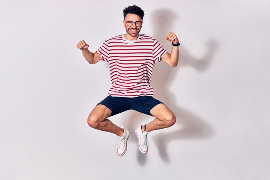 Young handsome man wearing casual clothes and glasses smiling happy. Jumping with smile on face doing winner sign with fists up over isolated white background