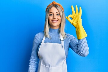 Beautiful blonde woman wearing cleaner apron and gloves smiling positive doing ok sign with hand...