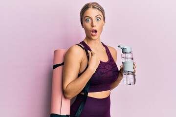Beautiful blonde woman wearing sportswear holding water bottle and yoga mat afraid and shocked with...