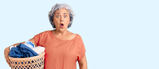 Senior woman with gray hair holding laundry basket scared and amazed with open mouth for surprise, disbelief face
