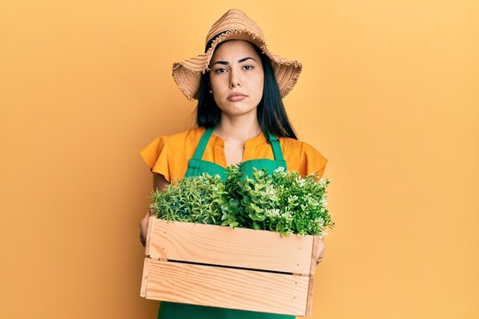 Beautiful young woman wearing gardener apron holding wooden plant pot relaxed with serious expression on face. simple and natural looking at the camera.