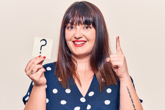 Young plus size woman holding question mark smiling with an idea or question pointing finger with happy face, number one