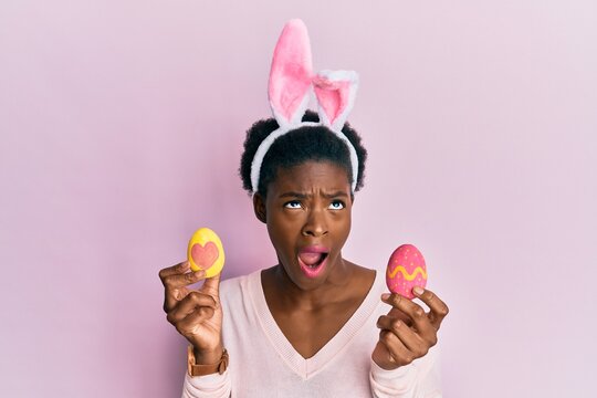 Young african american girl wearing cute easter bunny ears holding painted eggs angry and mad screaming frustrated and furious, shouting with anger looking up.
