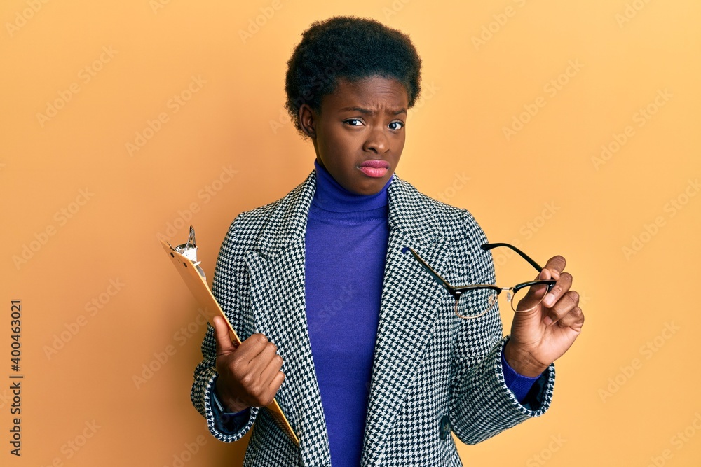 Poster Young african american girl holding clipboard and glasses clueless and confused expression. doubt concept.