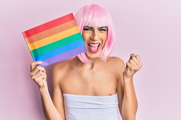 Young man wearing woman make up holding rainbow lgbtq flag wearing pink wig screaming proud,...