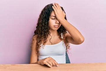 Teenager hispanic girl wearing casual clothes sitting on the table surprised with hand on head for...