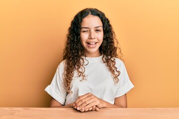 Teenager hispanic girl wearing casual clothes sitting on the table winking looking at the camera...