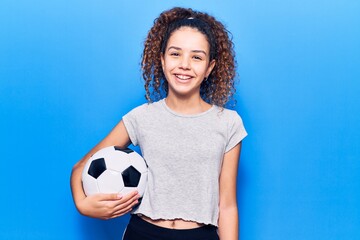 Beautiful kid girl with curly hair holding soccer ball looking positive and happy standing and...