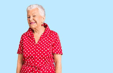 Senior beautiful woman with blue eyes and grey hair wearing a red summer dress winking looking at...