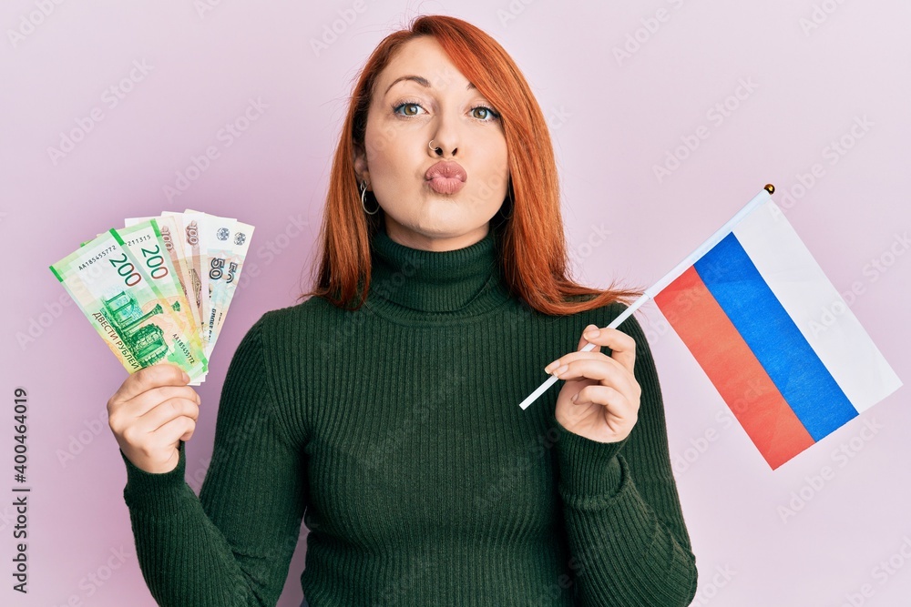 Sticker Beautiful redhead woman holding russian ruble banknotes and russia flag looking at the camera blowing a kiss being lovely and sexy. love expression.