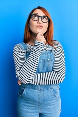 Beautiful redhead woman wearing casual clothes and glasses thinking concentrated about doubt with finger on chin and looking up wondering