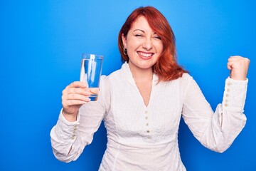 Young beautiful redhead woman drinking glass of water over isolated blue background screaming proud, celebrating victory and success very excited with raised arm