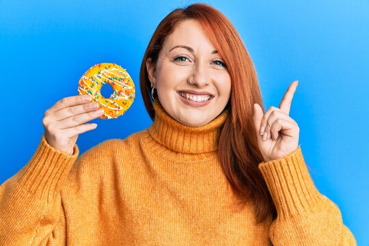 Beautiful redhead woman holding tasty colorful doughnut smiling happy pointing with hand and finger to the side