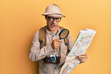 Middle age bald man wearing explorer hat holding magnifying glass on a map winking looking at the...