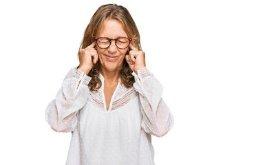 Middle age blonde woman wearing casual white shirt and glasses covering ears with fingers with annoyed expression for the noise of loud music. deaf concept.