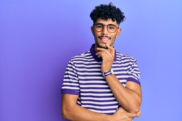 Fototapeta na wymiar Young arab handsome man wearing casual clothes and glasses smiling looking confident at the camera with crossed arms and hand on chin. thinking positive.