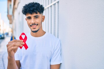 Young arab man smiling happy holding hiv awaraness red ribbon leaning on the wall.