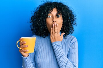 Young hispanic woman holding coffee covering mouth with hand, shocked and afraid for mistake....