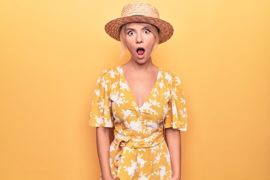 Beautiful blonde woman on vacation wearing summer hat and dress over yellow background afraid and shocked with surprise and amazed expression, fear and excited face.