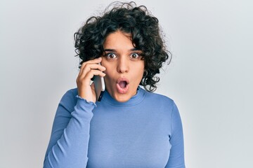 Young hispanic woman with curly hair having conversation talking on the smartphone scared and amazed with open mouth for surprise, disbelief face