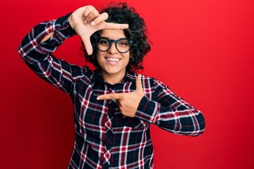 Fototapeta premium Young hispanic woman with curly hair wearing casual clothes and glasses smiling making frame with hands and fingers with happy face. creativity and photography concept.