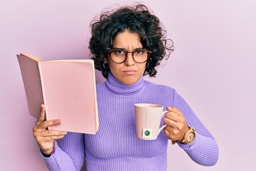 Young hispanic woman with curly hair reading a book and drinking a cup of coffee skeptic and nervous, frowning upset because of problem. negative person.