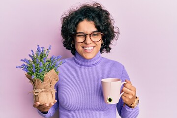 Young hispanic woman with curly hair drinking a cup of infused lavender winking looking at the...