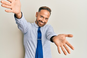 Handsome middle age man wearing business shirt and tie looking at the camera smiling with open arms for hug. cheerful expression embracing happiness.