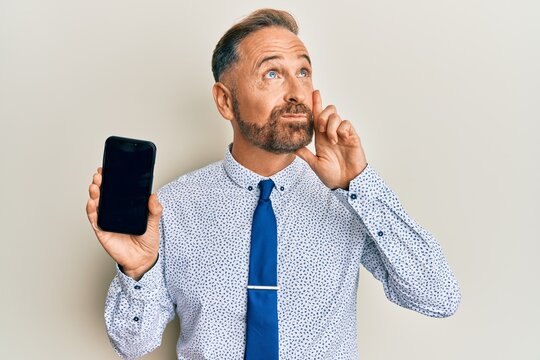 Handsome middle age business man holding smartphone showing blank screen serious face thinking about question with hand on chin, thoughtful about confusing idea