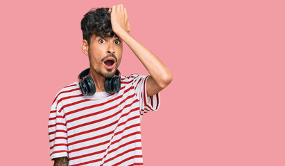 Young hispanic man listening to music using headphones surprised with hand on head for mistake,...