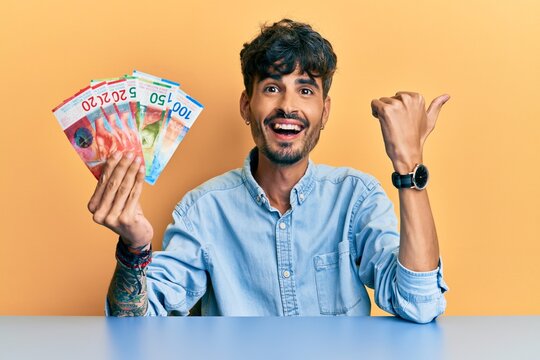Young hispanic man holding swiss franc banknotes sitting on the table pointing thumb up to the side smiling happy with open mouth