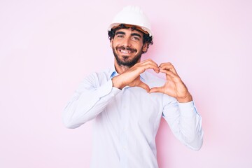 Handsome young man with curly hair and bear wearing architect hardhat smiling in love doing heart symbol shape with hands. romantic concept.