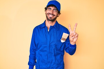 Handsome young man with curly hair and bear wearing builder jumpsuit uniform smiling with happy face winking at the camera doing victory sign. number two.