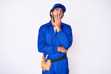 Handsome young man with curly hair and bear weaing handyman uniform bored yawning tired covering mouth with hand. restless and sleepiness.