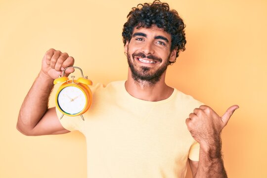 Handsome young man with curly hair and bear holding alarm clock pointing thumb up to the side smiling happy with open mouth